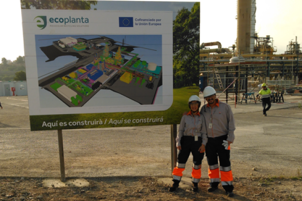 Two people are standing on a factory site in front of a sign containing the floor plan
