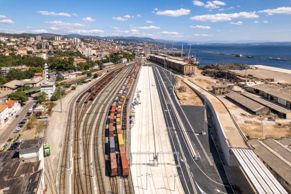 Image of Rijeka port showing the 4 new rail tracks with the blue sea in the background. There are several rail carriages on the rails in the colours of yellow, red and blue.